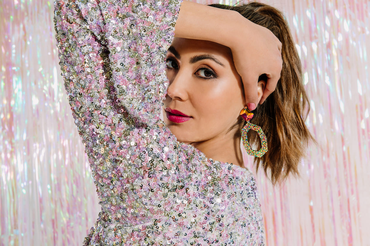 Peering behind left arm with Jaci crystal glitter earrings on left ear against iridescent streamer wall background
